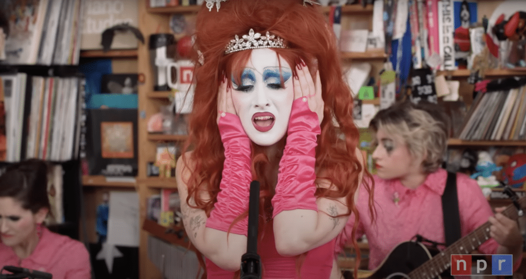 Chappell Roan, wearing pale foundation with massive blue eyeshadow, sings in front of a messy desk.