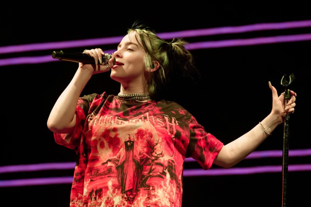 Billie Eilish onstage singing into a microphone, wearing red tour merch.