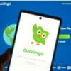 A phone is held up, showing the Duolingo mascot waving.