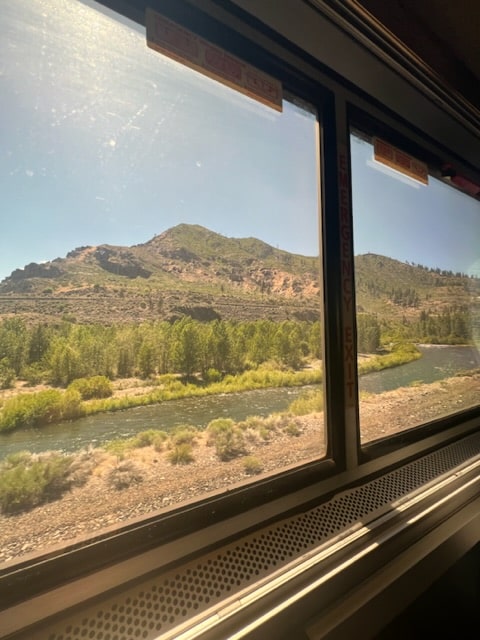 Green topped mountains over the Colorado River through train window. River is subtle green, under open blue sky/