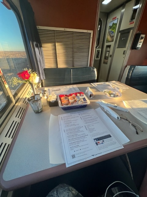 Dinner car. Menu, silverware, tiny box holding honey, James, ketchup, creamer, a vase with a white and red rose.