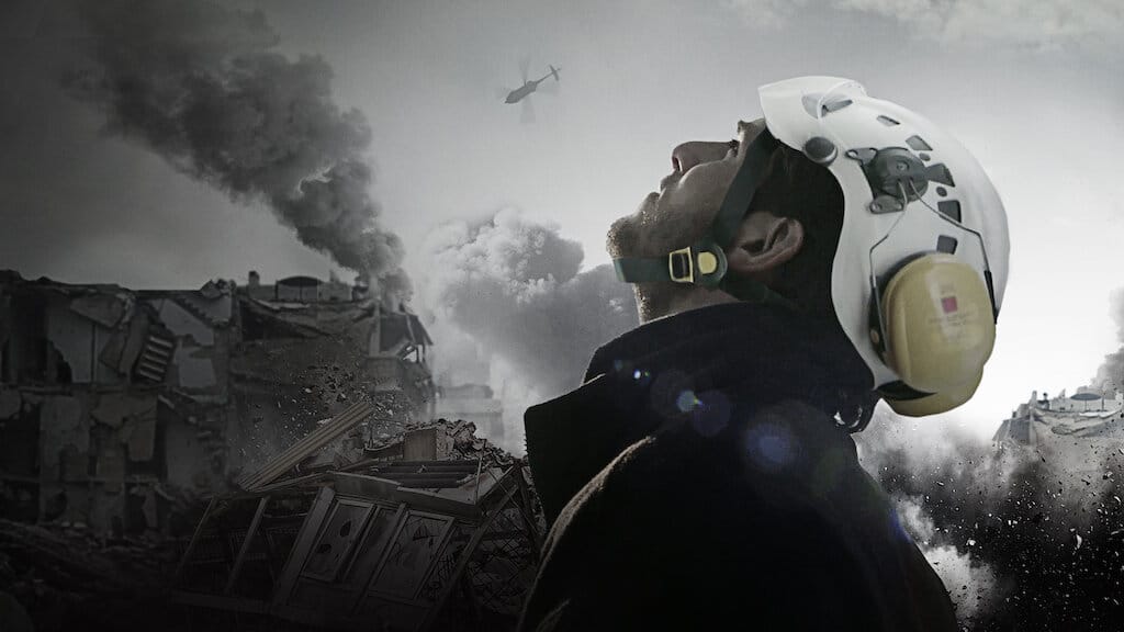 A White Helmet looks toward the smokey sky with a building on fire in the background.