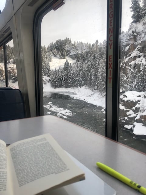 A book and pen next to train window, viewing the Colorado River in the winter, almost black, and snow capped trees.