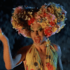 A man in drag, with thin, drawn-on eyebrows, blue eyeshadow, and red lipstick. They have a floral crown on their head. Hugo Weaving as Mitzi Del Bra in 'Priscilla, Quen of the Desert' performing a drag routine