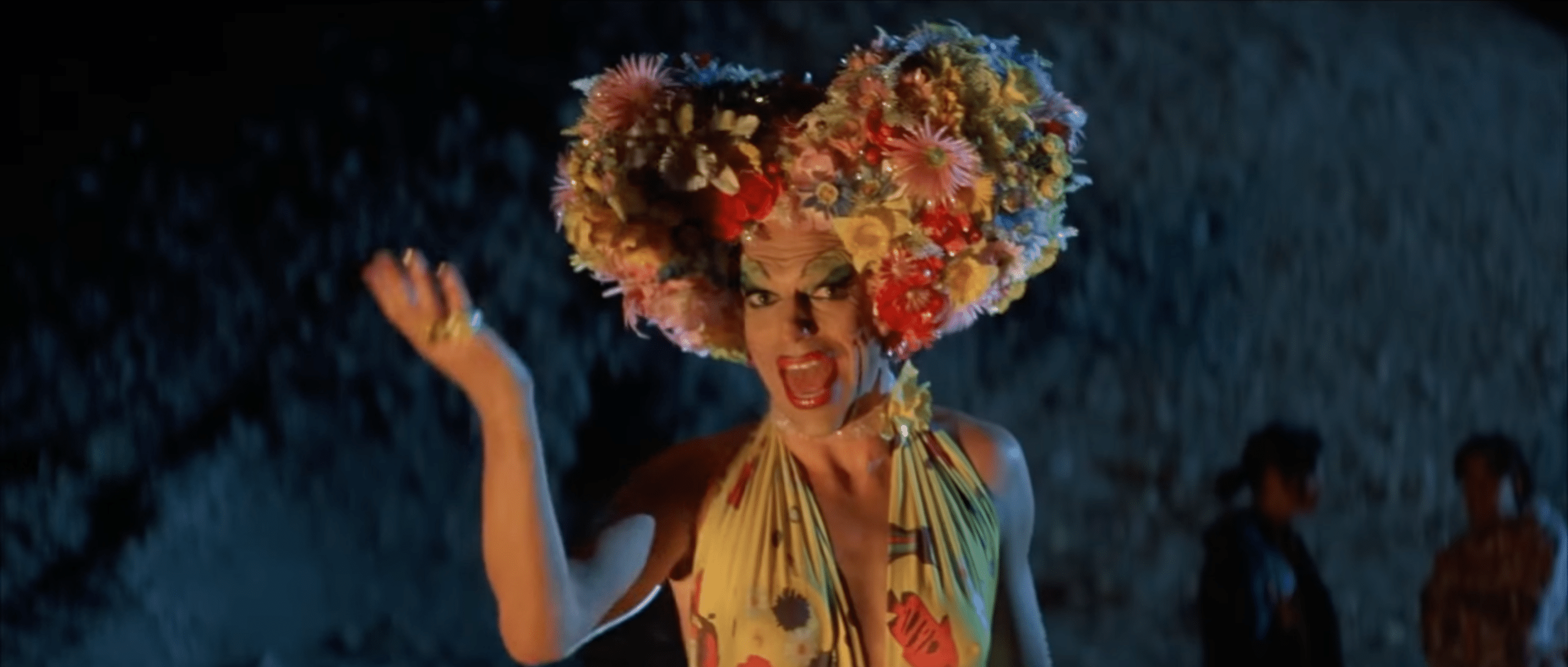 A man in drag, with thin, drawn-on eyebrows, blue eyeshadow, and red lipstick. They have a floral crown on their head. Hugo Weaving as Mitzi Del Bra in 'Priscilla, Quen of the Desert' performing a drag routine