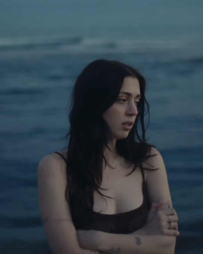 An image of Lizzy McAlpine with the blue background of a lake behind her, from her music video.