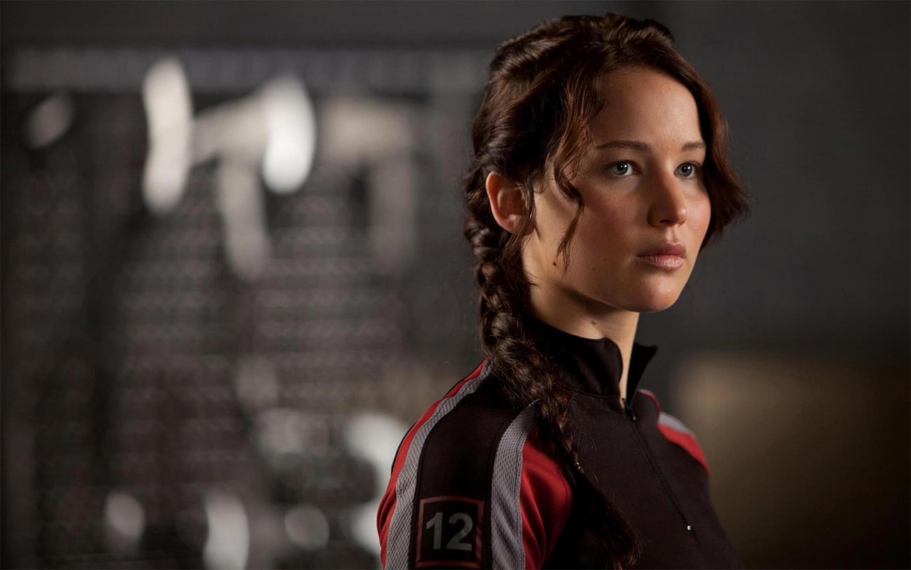 Katniss Everdeen stands in front of a wall of blades and axes.