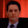 Kyle MacLachlan as Dale Cooper in Twin Peaks. A man with dark hair in a dark suit sat down. He is backgrounded by a red curtain.