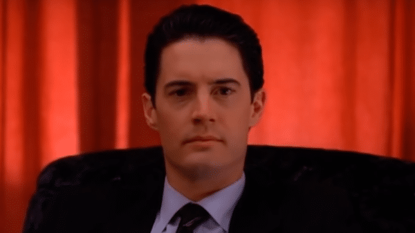Kyle MacLachlan as Dale Cooper in Twin Peaks. A man with dark hair in a dark suit sat down. He is backgrounded by a red curtain.