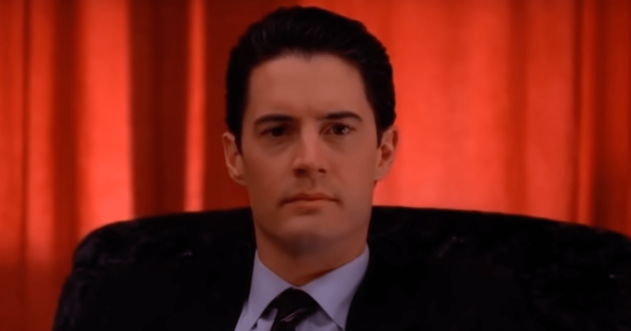 Kyle MacLachlan as Dale Cooper in Twin Peaks. A man with dark hair in a dark suit sat down. He is backgrounded by a red curtain.