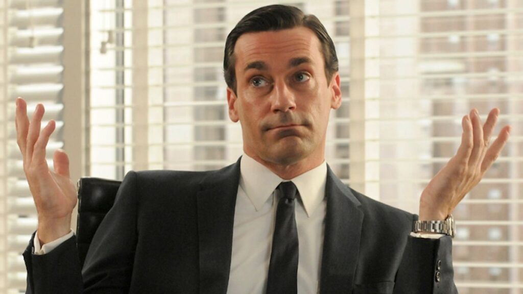 Jon Hamm as Don Draper in 'Mad Men'. A man in a suit, throwing his hands up indifferently. He is in the office of a high rise.