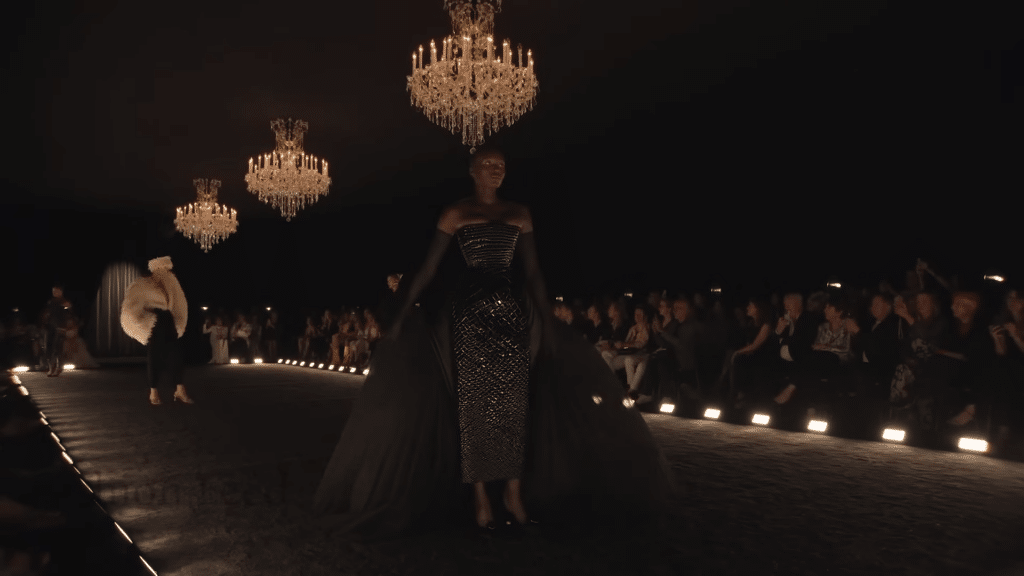 Runway model sporting a long black butterfly-style dress with black resin sequins and black gloves.