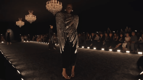 Runway model showcasing a black cape with chromed faux feather adornment.
