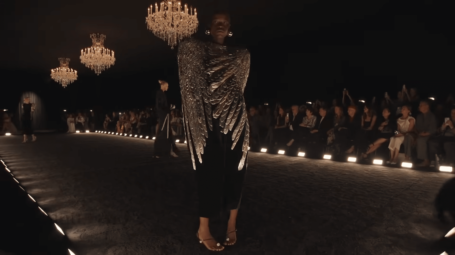 Runway model showcasing a black cape with chromed faux feather adornment.