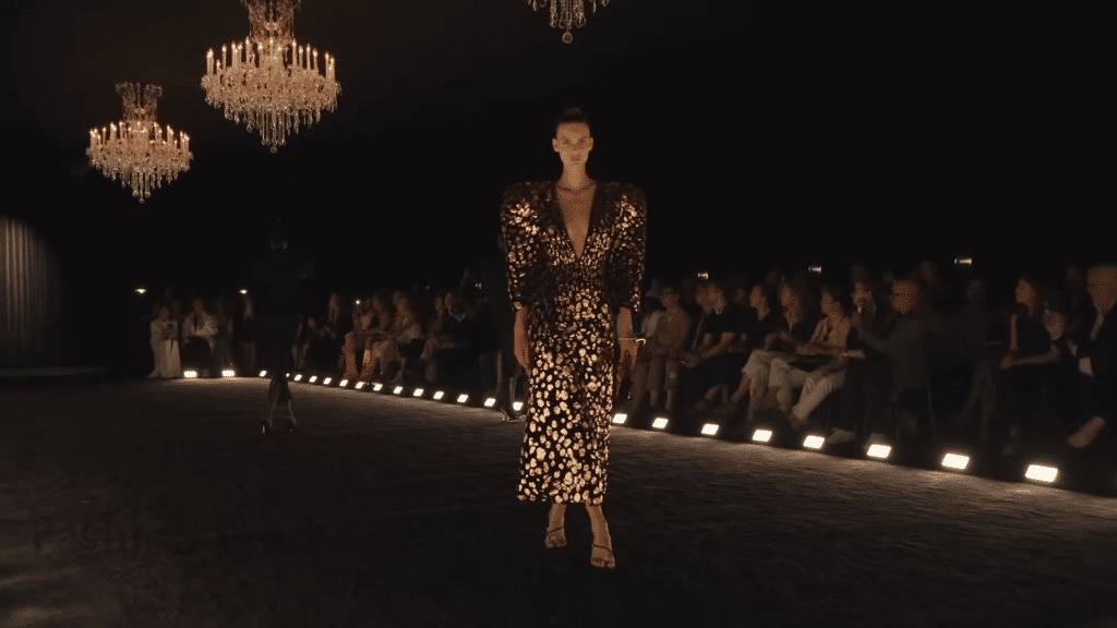 Runway model sporting a long gold and black dress with accentuated shoulders.