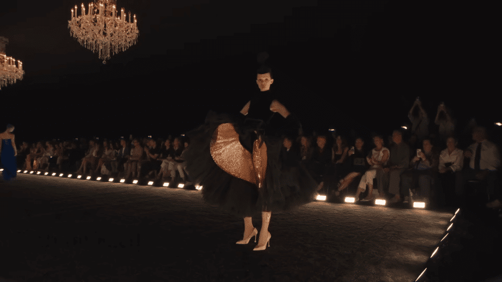 Runway model sporting a black dress with an upturned "kick" that is accentuated by pink rhinestones.