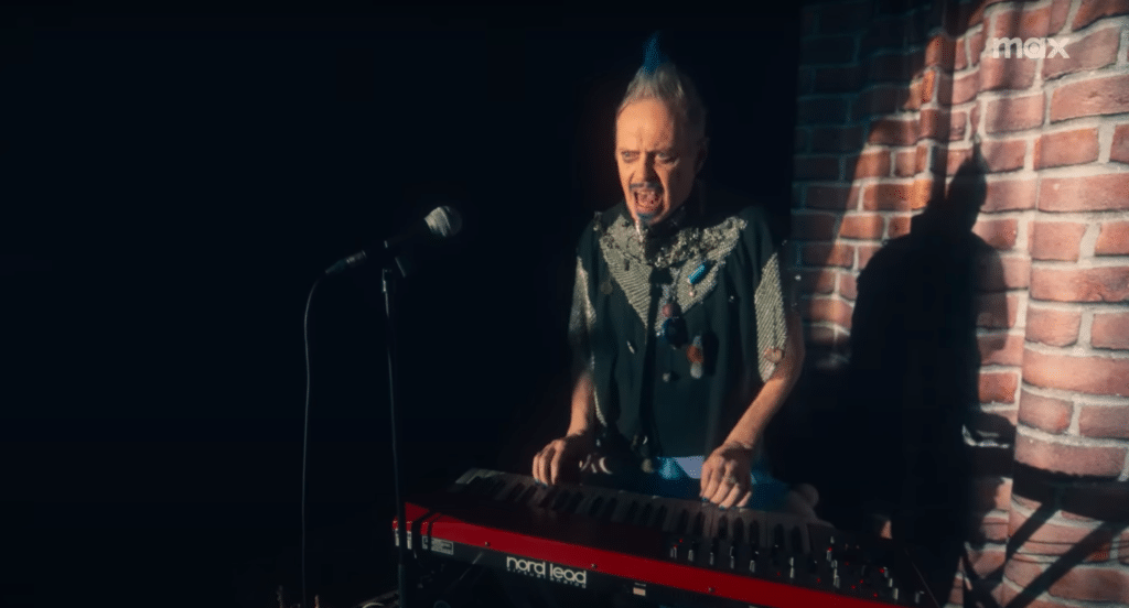 Steve Buscemi, as the character Q, is behind a keyboard and microphone, preparing to perform at an open mic night.