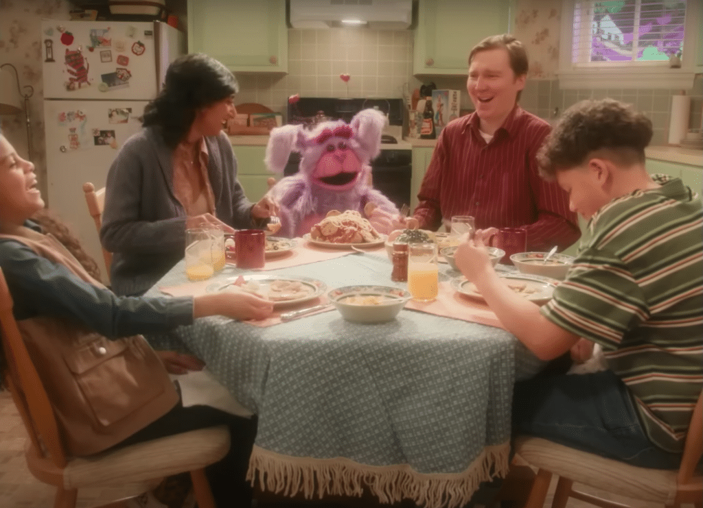 A family is sitting at a table, having dinner and laughing. At the center, is a large purple puppet.