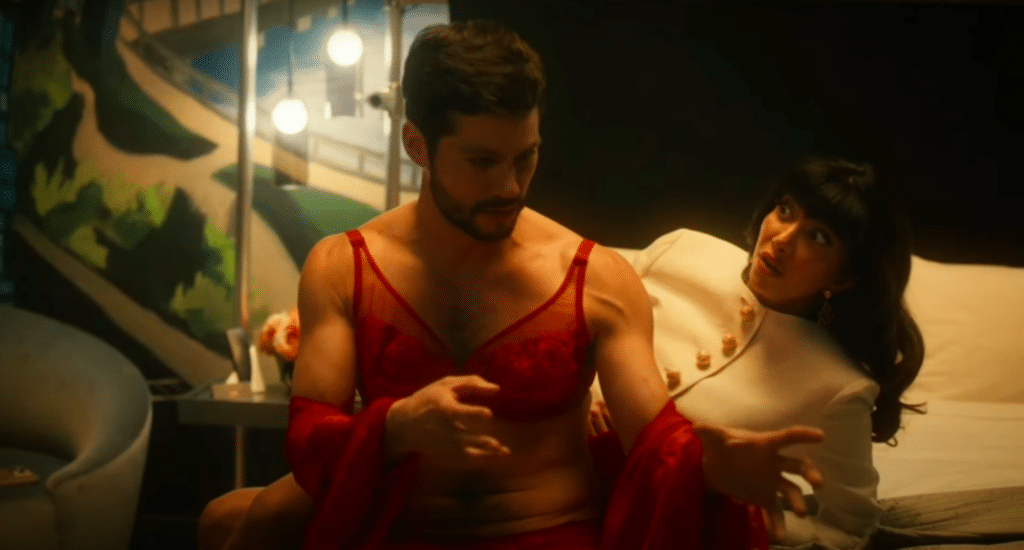 Dylan O'Brien is sitting on a bed while wearing red lingerie. Behind him is a woman lying down in a white dress. She is watching him with a slightly disgusted expression, unrelated to the lingerie.