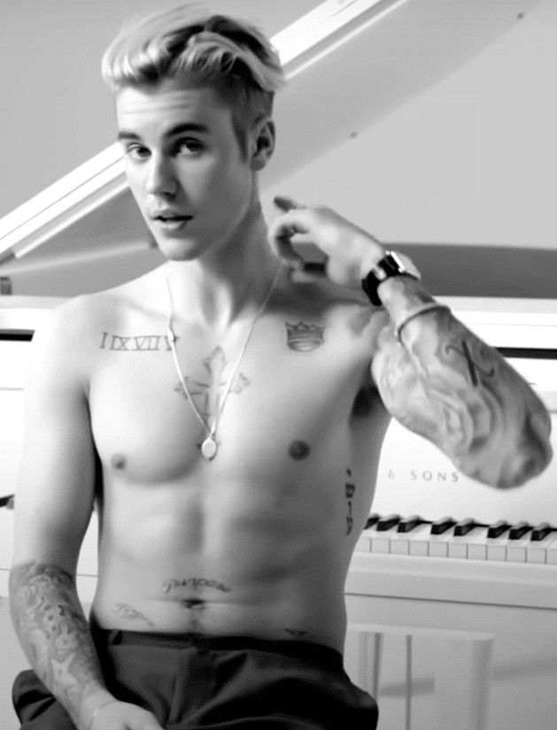 Justin Bieber showing off his tattoos while sitting in front of a piano.