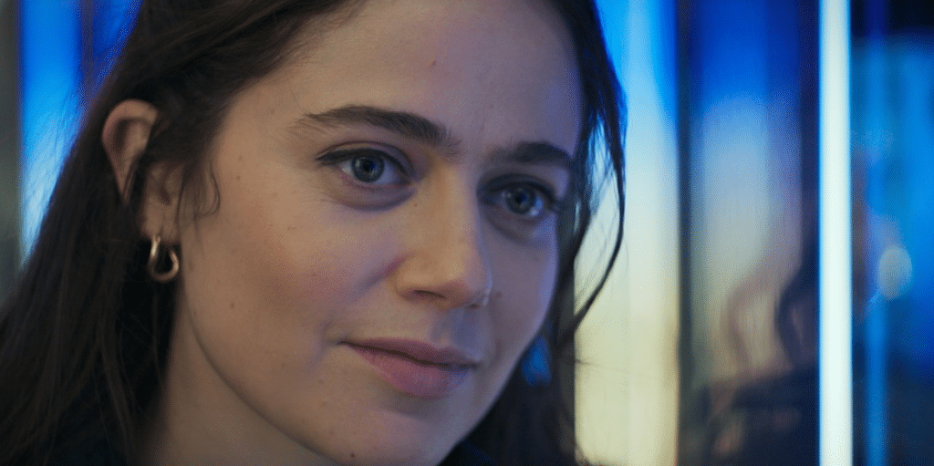 A close-up of a young woman with long, dark hair and blue eyes, wearing gold hoop earrings. She has a slight smile on her face and is illuminated by soft, blue-tinted lighting in the background, giving the image a calm and introspective atmosphere.