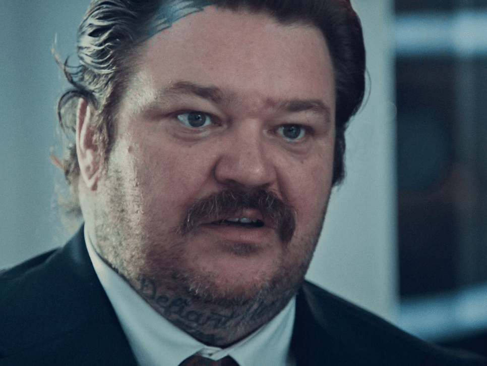 A man with a moustache and tattoos on his forehead, stares straight ahead looking slightly confused.