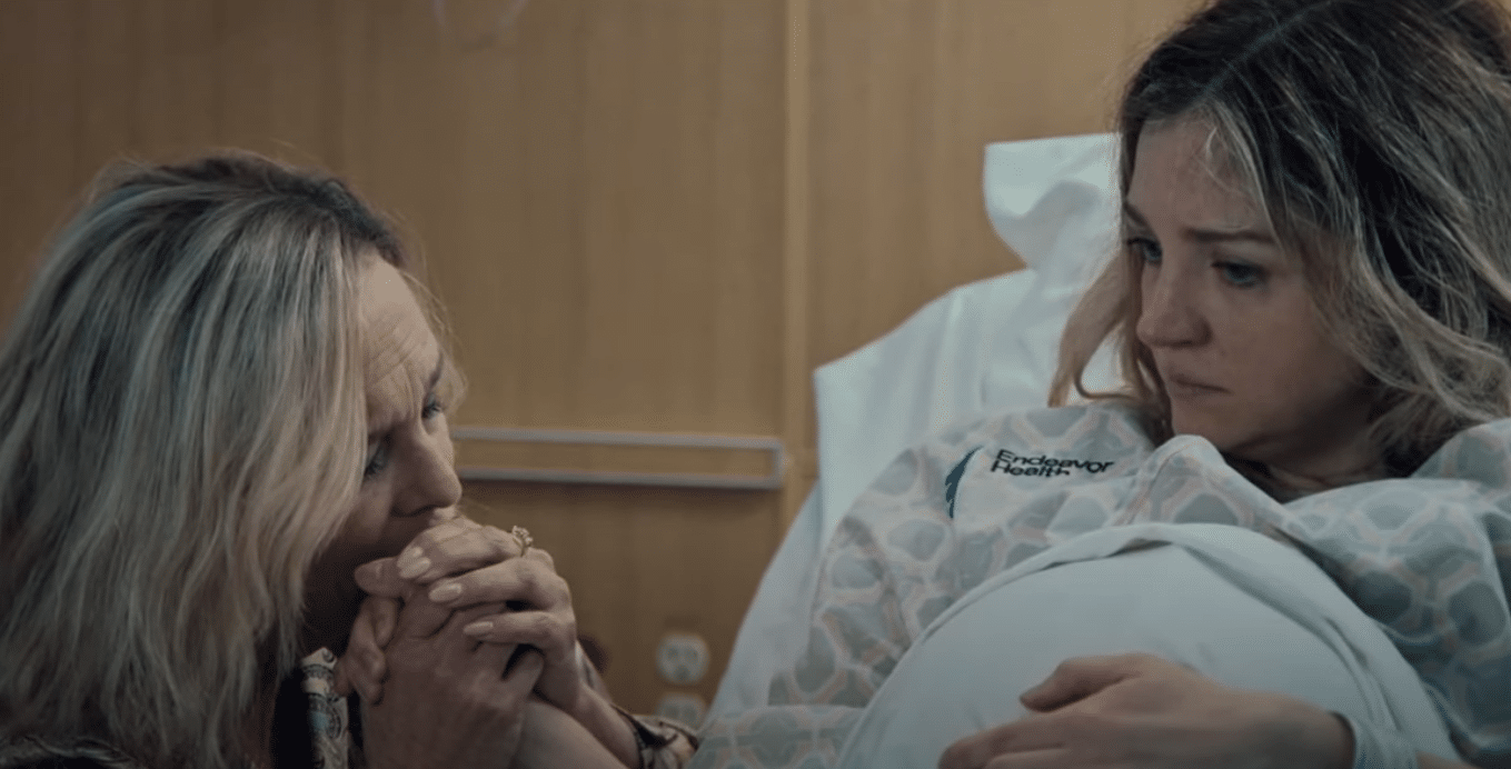A woman in a hospital gown sits in a bed, looking down with a concerned expression. Another woman, sitting beside her, clasps her hands tightly and brings them to her lips, showing support and emotional connection. The setting appears to be a hospital room, adding to the scene's emotional intensity.