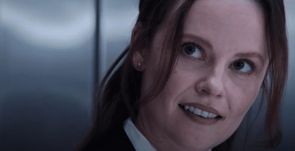 A close-up of a woman with brown hair, partially tied back, and a few loose strands framing her face. She is smiling slightly and looking to the side. She is wearing a dark jacket and a white shirt, with a metallic, blurred background behind her.