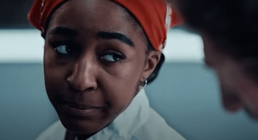 A close-up of a woman wearing a red headscarf and white chef's uniform. She has a serious, contemplative expression on her face, looking slightly to the side. Another person is partially visible in the foreground, blurred.