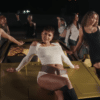 A group of young women are posing around a gold car at night. The woman in the center is sitting on the hood, wearing a white crop top and shorts with a wide belt. The woman on the left is leaning against the car, wearing a white crop top and blue jeans. The woman on the right is leaning on the car, wearing a black dress and brown boots. Other women are visible in the background, adding to the dynamic and stylish scene.