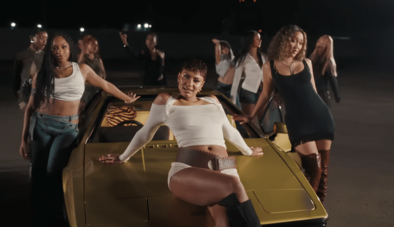 A group of young women are posing around a gold car at night. The woman in the center is sitting on the hood, wearing a white crop top and shorts with a wide belt. The woman on the left is leaning against the car, wearing a white crop top and blue jeans. The woman on the right is leaning on the car, wearing a black dress and brown boots. Other women are visible in the background, adding to the dynamic and stylish scene.