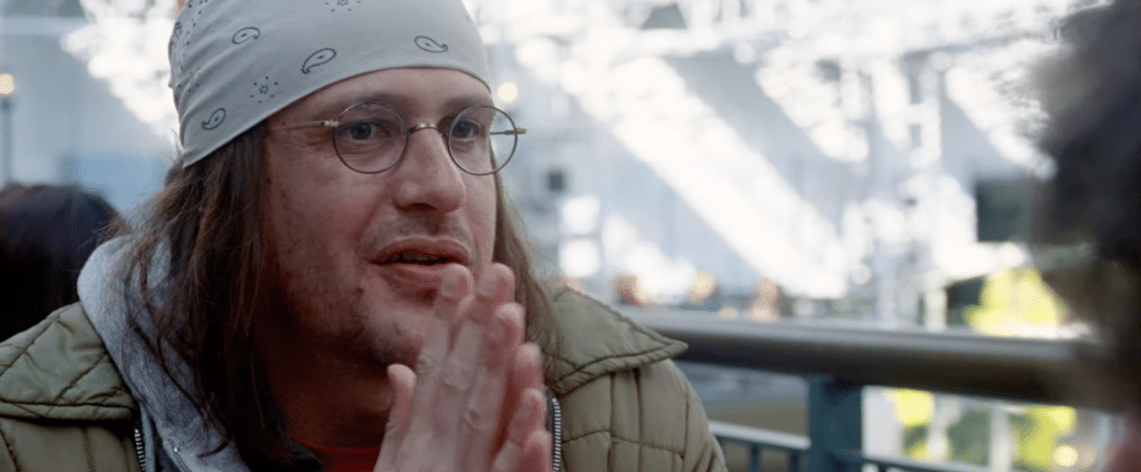 Jason Segel as David Foster Wallace. A man with round glasses and a white bandana. He is speaking, holding his hands in front of his face.