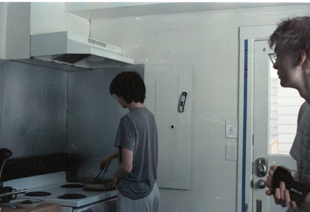 Two people are standing in a kitchen; one is cooking at the stove and the other is standing behind, closer to the camera. The image has a lot of scratches on it.