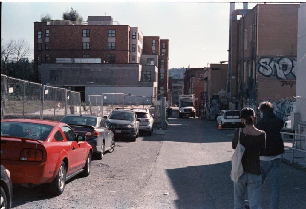 Two people are walking through an alleyway. Next to them is a line of cars. This image has more magenta to it than the one before.