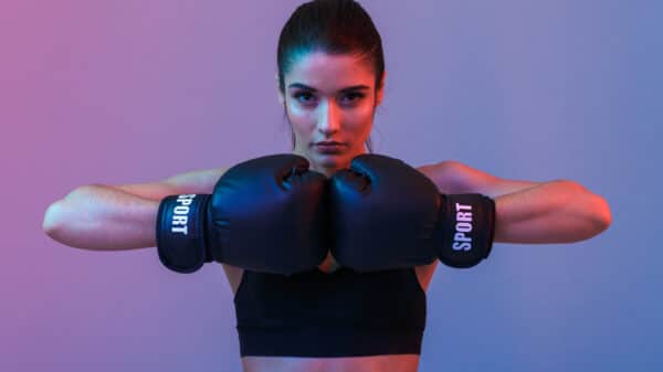 Image of young sports woman in tracksuit and black putting hands together in boxing gloves isolated over purple background