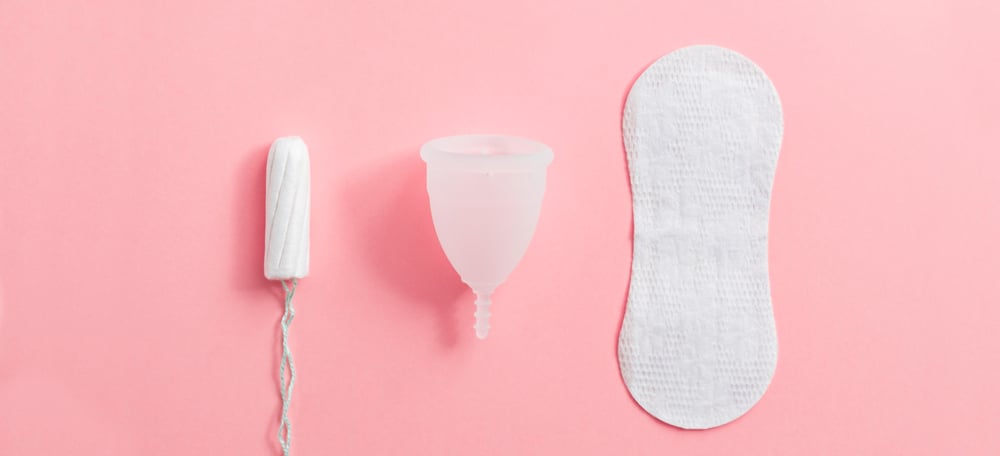 A tampon, menstrual cup, and pad lying in a row. Light pink background.