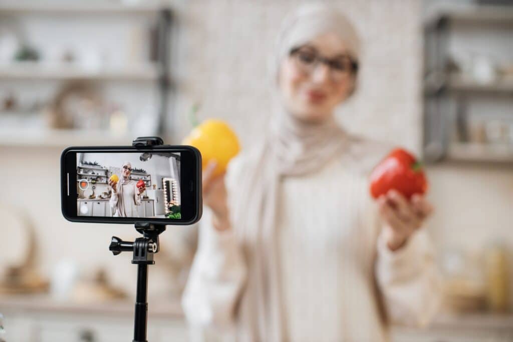 Woman holding bell peppers in each hand while posing in front of her phone.