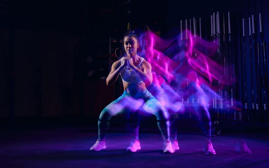 Squats. Sportive woman in sportswear and sportsfoot training in modern fitness room over dark background with mixed neon light. Active and healthy lifestyle concept