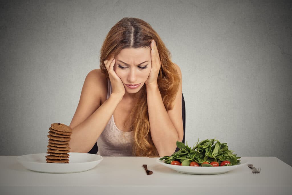 Woman sitting at a table in front of a grey background. She is sitting between a plate of salad and a plate of cookies and looks at the cookies with a frustrated expression.