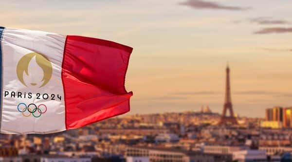 A French flag branding the 2024 Olympics' logo flies in the forefront of an out-of-focus photo of Paris and the Eiffel Tower.