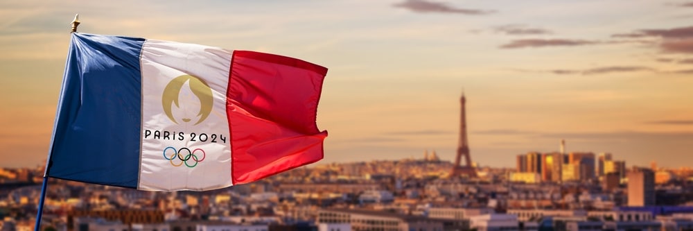 A French flag branding the 2024 Olympics' logo flies in the forefront of an out-of-focus photo of Paris and the Eiffel Tower.
