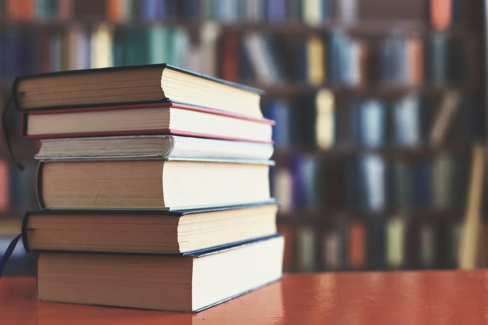 photo of stack of books with the pages facing the camera. the books are on a reddish wooden table and blurred in the background are bookshelves filled with books