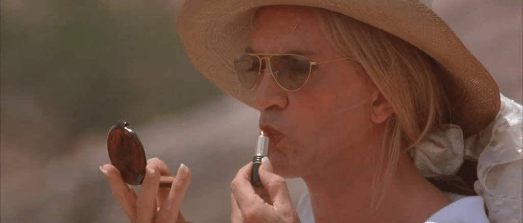 A woman applying lipstick. She is in a sunhat and a white shirt. Terence Stamp as Bernadette in 'Priscilla, Queen of the Desert'