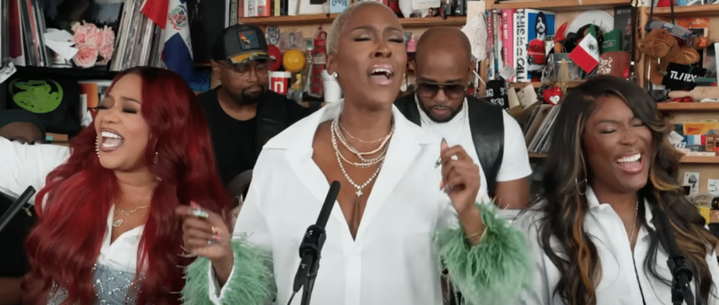 The R&B girl group SWV is performing in an intimate setting for a Tiny Desk concert. The three members, dressed in stylish white outfits with green feathered cuffs, are passionately singing with their hands raised. Behind them, musicians are playing instruments, and shelves filled with books and various items create a cozy backdrop.