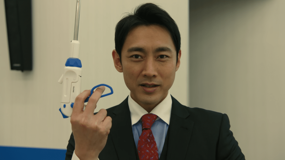 Takashina portrayed by Kotaro Koizumi holds up a white medical device that is gun shaped