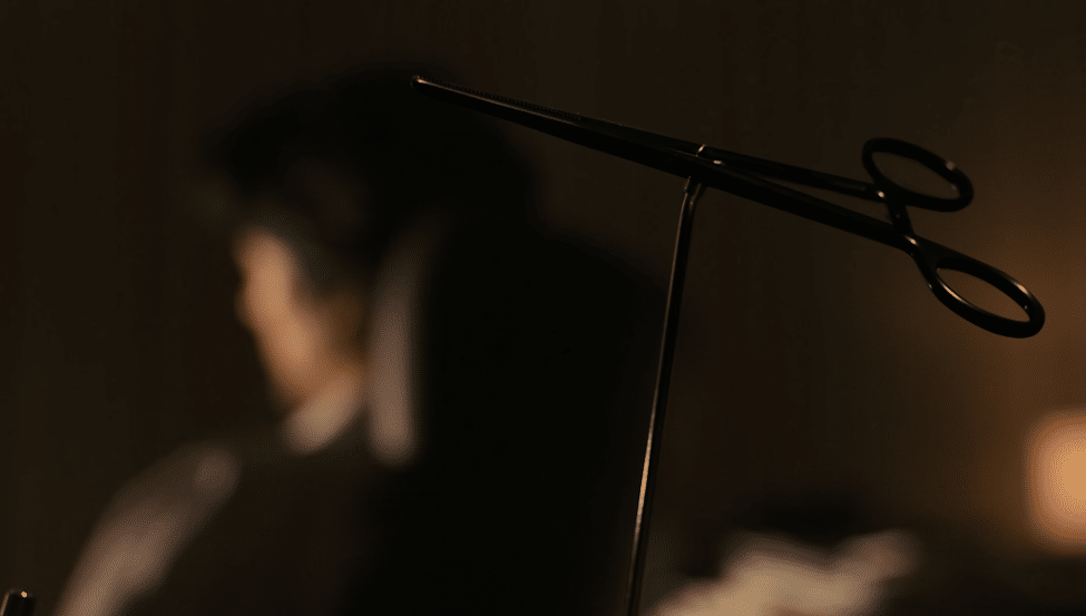 In focus a pair of black forceps, out of focus in the background is Dr Saeki facing away