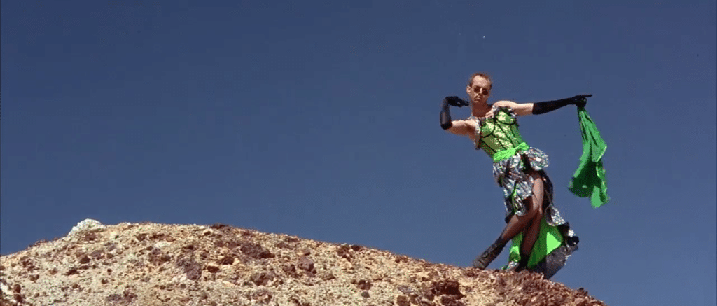A man in drag at the top of a rock. He is dressed in a green and black dress and is holding his arms out to his left. Hugo Weaving as Tick/Mitzi in 'Priscilla, Queen of the Desert'