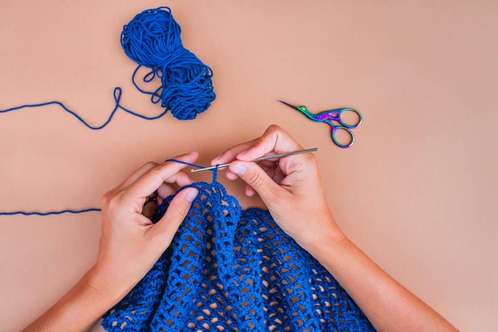 view of hands crocheting blue yarn on beige background