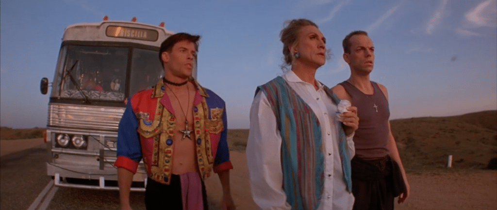 Two men and a woman in front of a bus in the middle of the road. One man is in a colourful jacket, the other in a brown vest. The woman is wearing a white shirt and a denim vest. Pearce, Stamp, and Weaving in 'Priscilla, Queen of the Desert'