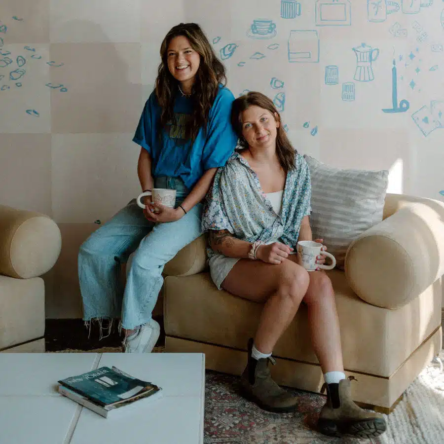 two girls sit on a couch holding coffee mugs